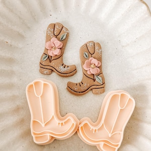 May include: Two decorated cowboy boot cookies with pink flower accents and two pink cowboy boot-shaped cookie cutters. The cookies are light brown with silver glitter details. The cutters are a light peach color.