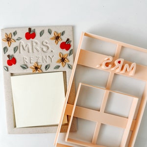 May include: A beige and brown clay sign with a cutout for a sticky note. The sign is decorated with fall-themed embellishments, including leaves, flowers, and apples. The sign also has the text "MRS. REZNY" embossed on it. A separate plastic mold is also shown, which is used to create the sign. The mold has a cutout for the text "MRS."