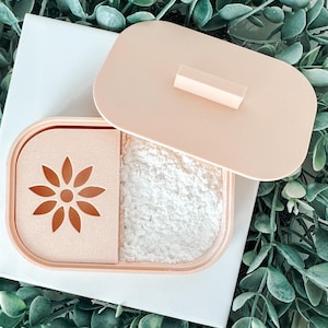 May include: A light peach-colored container with a lid, holding white powder. The container has a compartment with a flower-shaped cutout. The lid has a small rectangular handle. The container is on a white surface, surrounded by green foliage.