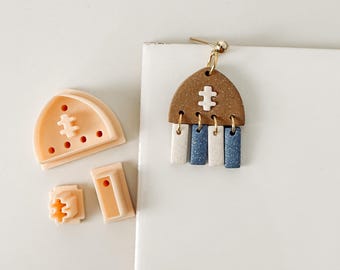 May include: A handmade earring with a brown arch-shaped top and three dangling rectangular pieces in white and blue. The earring has a gold-colored post and a small white accent on the top. Beside the earring are clay cutters in the same shape and design.