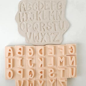 May include: A beige alphabet stamp set with a matching letter mold. The mold features the letters A through Z in a sans-serif font. The individual stamps are arranged in a grid, each with a single letter. The set is ideal for crafting and educational projects.
