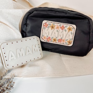 May include: A white ceramic plaque with the word "AMAM" in raised letters, and a black zippered pouch with a white patch that says "MAMA" in raised letters and is decorated with embroidered flowers.