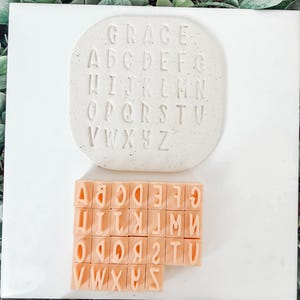 May include: A set of alphabet stamps and a matching impression block. The impression block is off-white with the word "GRACE" and the alphabet imprinted. The stamps are peach-colored and arranged in rows, each with a letter.