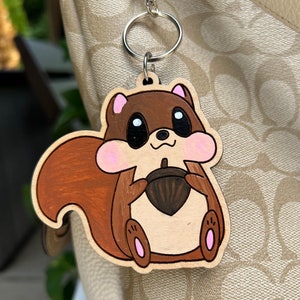 May include: A wooden keychain shaped like a cartoon squirrel holding an acorn. The squirrel is brown with pink cheeks and a white belly. The acorn is brown and has a light brown cap.