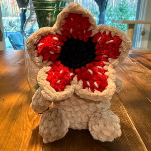 May include: A plush Demogorgon creature, featuring a beige body and a large, flower-like head with red and white detailing. The center of the head is black. The plush toy has small, rounded feet and is sitting on a wooden surface.