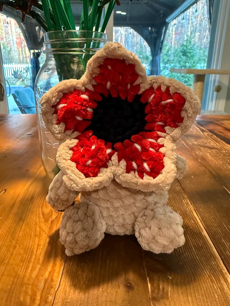 May include: A plush Demogorgon creature, featuring a beige body and a large, flower-like head with red and white detailing. The center of the head is black. The plush toy has small, rounded feet and is sitting on a wooden surface.