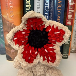 May include: A plush, beige Demogorgon-inspired creature with a red and white flower-like head and a black center. The soft toy is sitting in front of a shelf of books.