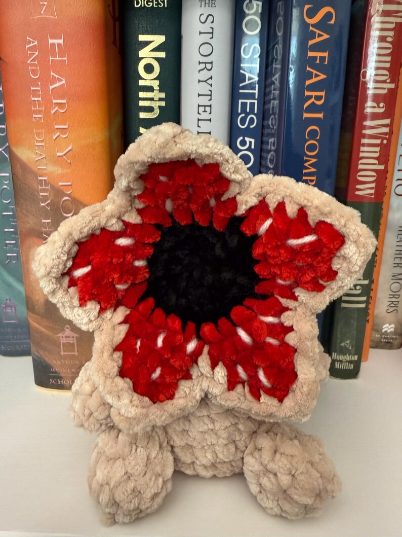 May include: A plush, beige Demogorgon-inspired creature with a red and white flower-like head and a black center. The soft toy is sitting in front of a shelf of books.