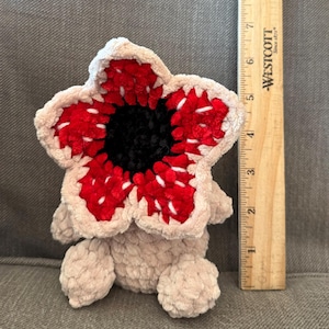 May include: A plush Demogorgon toy, featuring a flower-like head with red and white petal-like structures surrounding a black center. The body is a soft, light beige color. A wooden ruler is next to the toy, measuring approximately 4 inches in height.