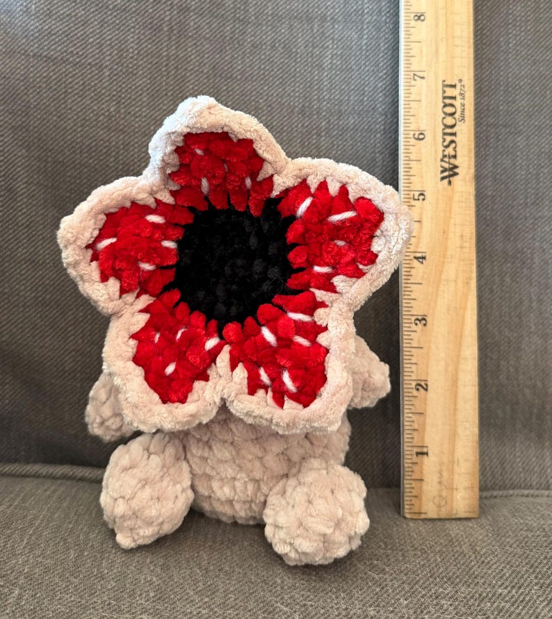May include: A plush Demogorgon toy, featuring a flower-like head with red and white petal-like structures surrounding a black center. The body is a soft, light beige color. A wooden ruler is next to the toy, measuring approximately 4 inches in height.