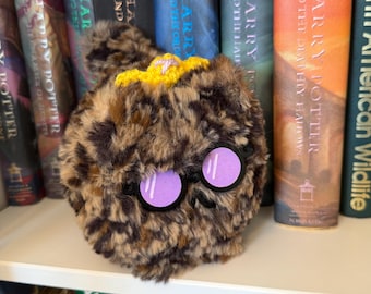 May include: A round, fuzzy, brown and black leopard-print plush toy featuring a small yellow crocheted crown on top and a pair of round black-rimmed glasses with purple lenses positioned on the front of the textured, soft material.