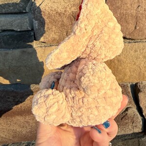 May include: A light peach-colored, plush, handmade crochet chicken toy with a red comb. The chicken is held in a hand against a stone wall background. The toy has a soft, textured appearance.