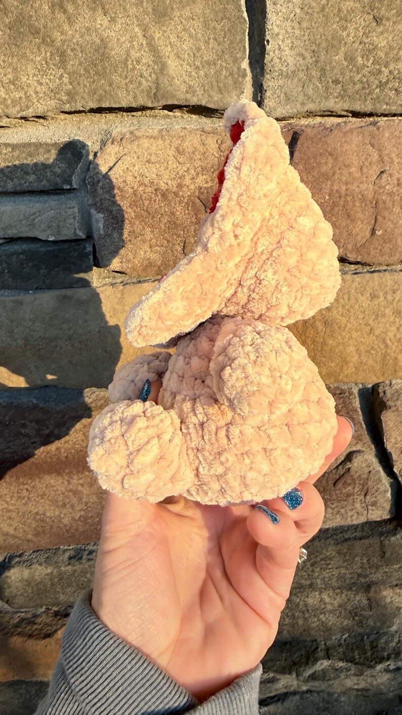 May include: A light peach-colored, plush, handmade crochet chicken toy with a red comb. The chicken is held in a hand against a stone wall background. The toy has a soft, textured appearance.