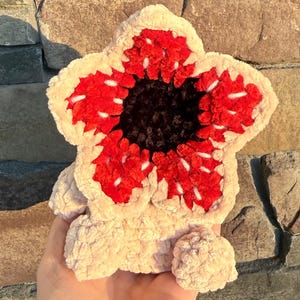 May include: A plush, cream-colored Demogorgon, a fictional monster from the series, with a red and black flower-like head. The creature has a textured, soft appearance, with a detailed, open-mouthed design. The background is a stone wall.