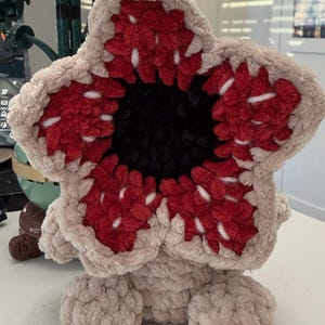 May include: A plush Demogorgon toy, featuring a beige body and a distinctive flower-like head. The head has red and white petal-like structures surrounding a black center. The toy is sitting on a white surface.