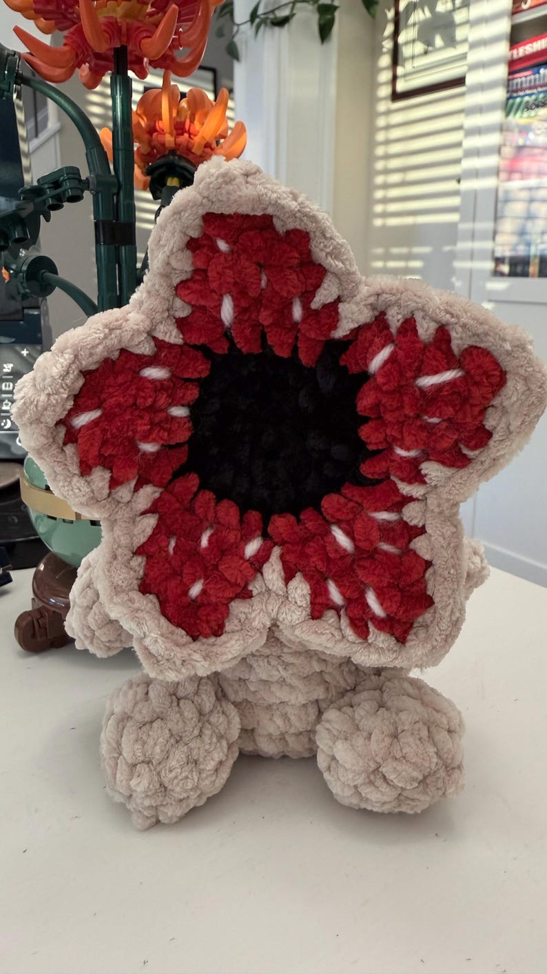 May include: A plush Demogorgon toy, featuring a beige body and a distinctive flower-like head. The head has red and white petal-like structures surrounding a black center. The toy is sitting on a white surface.