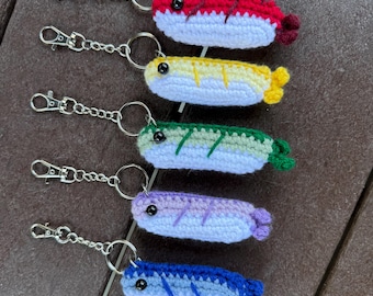 May include: Five crochet shrimp keychains with silver-tone metal lobster claw clasps and split rings. Each shrimp features a white underside, a colored top in red, yellow, green, purple, or blue, and a small black bead eye.