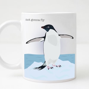 Peut inclure: Une tasse en céramique blanche avec une illustration en noir et blanc d'un pingouin sur un iceberg. Le texte "not gonna fly" est imprimé sur la tasse.