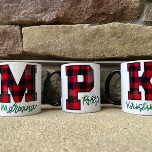 Pode incluir: Três canecas de cerâmica branca com alças pretas. Cada caneca apresenta uma grande letra em tecido xadrez vermelho e preto, "M", "P" e "K", com um nome por baixo. Os nomes são "Mariana", "Polly" e "Kristina".