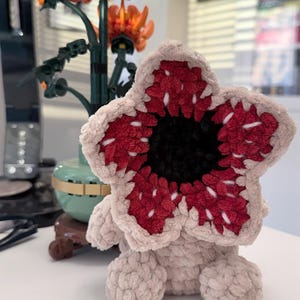 May include: A plush Demogorgon creature, featuring a beige body and a large, open flower-like head with red and white detailing. The center of the head is black. The toy is sitting on a white surface.