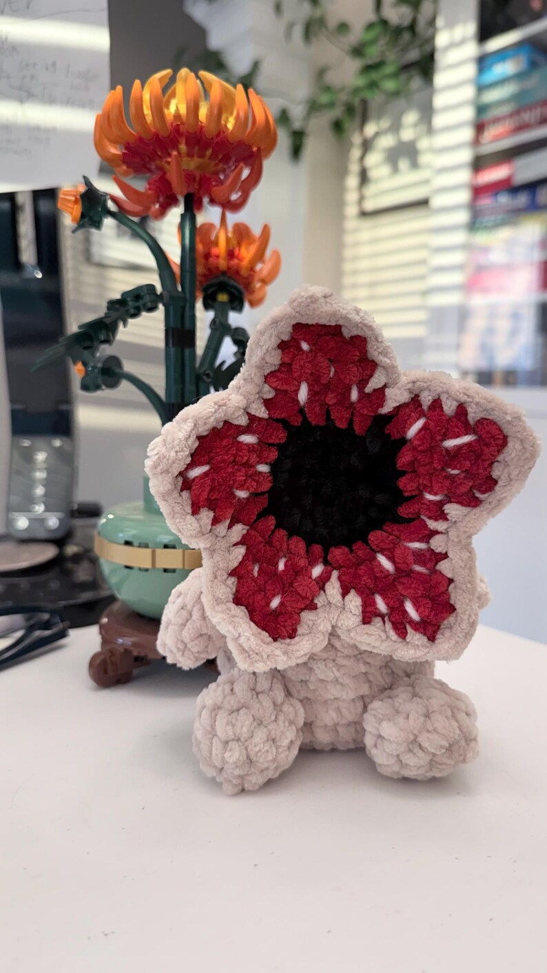 May include: A plush Demogorgon creature, featuring a beige body and a large, open flower-like head with red and white detailing. The center of the head is black. The toy is sitting on a white surface.