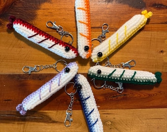 May include: Six crocheted shrimp keychains arranged in a circle. Each keychain features a white body with a colored spine and tail, including red, orange, yellow, green, blue, and purple variations. Each shrimp has a black bead eye and a silver metal lobster clasp attached by a chain.