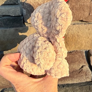 May include: A light pink, plush bunny toy with a textured surface. The bunny has a red accent and is held in a hand. The background is a stone wall.