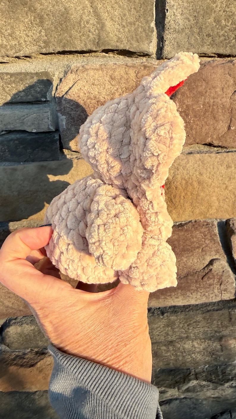 May include: A light pink, plush bunny toy with a textured surface. The bunny has a red accent and is held in a hand. The background is a stone wall.