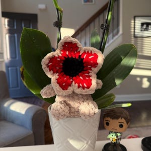 May include: A handmade crochet Demogorgon flower, featuring a red and white flower head with a black center, and a tan body. The Demogorgon is placed in a white patterned pot with green leaves. A Star Wars figurine is next to the pot.