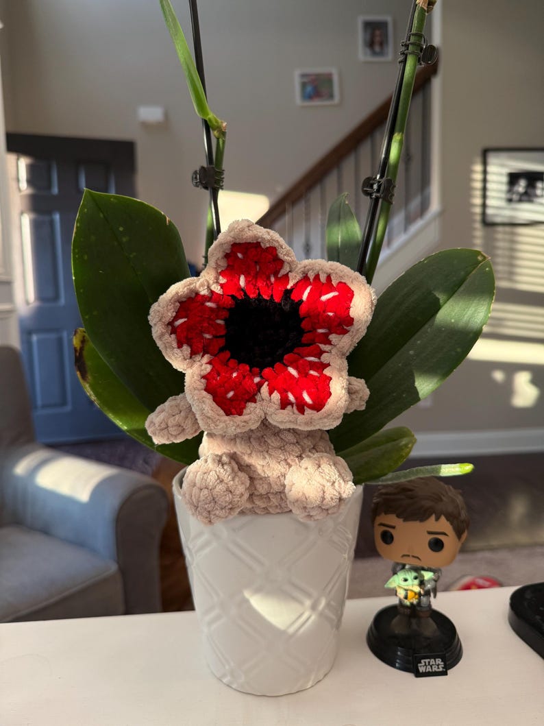 May include: A handmade crochet Demogorgon flower, featuring a red and white flower head with a black center, and a tan body. The Demogorgon is placed in a white patterned pot with green leaves. A Star Wars figurine is next to the pot.
