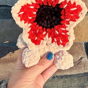 May include: A plush, Demogorgon-inspired creature, featuring a cream-colored, petal-like exterior with a vibrant red and black center. The handmade plush has a soft, textured appearance, with white accents and small, rounded feet.
