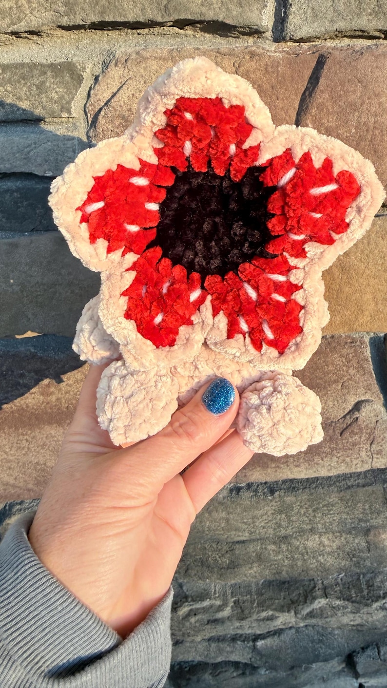 May include: A plush, Demogorgon-inspired creature, featuring a cream-colored, petal-like exterior with a vibrant red and black center. The handmade plush has a soft, textured appearance, with white accents and small, rounded feet.