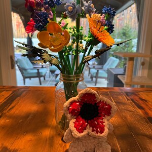 May include: A bouquet of colorful artificial flowers in a clear glass jar on a wooden table. The flowers include orange, blue, and purple blooms. A crocheted Demogorgon plush toy sits in front of the jar.