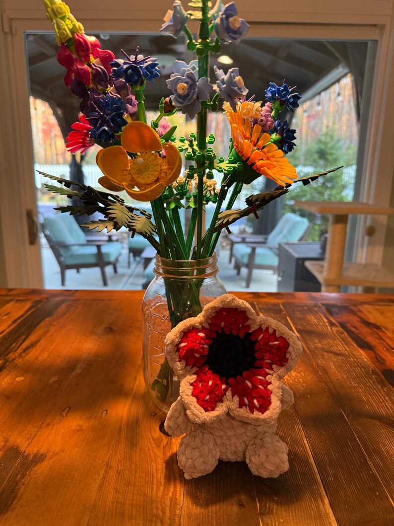 May include: A bouquet of colorful artificial flowers in a clear glass jar on a wooden table. The flowers include orange, blue, and purple blooms. A crocheted Demogorgon plush toy sits in front of the jar.