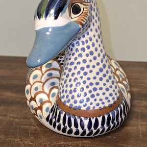 May include: A ceramic duck figurine featuring a white base with hand-painted blue polka dots on the neck and head. The wings display a pattern of brown and blue scalloped shapes, while the base is accented with dark blue vertical brushstrokes.