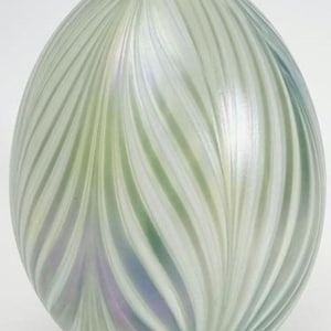 May include: A decorative, oval-shaped glass object with a swirling pattern of green, white, and iridescent colors. The design features vertical, curved lines that create a textured surface. The overall shape is smooth and rounded.
