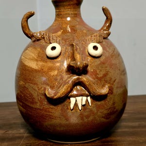 May include: A round, brown glazed ceramic jug featuring a sculpted face with a prominent nose, a wide mustache, and three white teeth. The vessel has two curved horn-like handles on the sides, two round white eyes, and textured eyebrows.