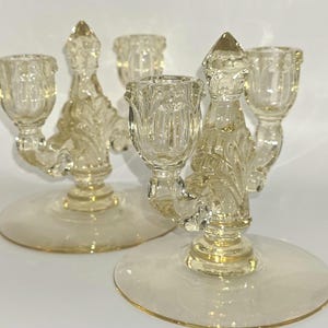 May include: Two clear glass candelabras with a decorative design. Each candelabra has three candle holders and a round base. The glass has a slight yellow tint, and the design includes floral and leaf patterns.