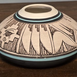May include: A low-profile, rounded ceramic vessel featuring a cream-colored upper body with etched black geometric patterns and stylized feather motifs. A thin turquoise band encircles the neck and lower section, which transitions to a solid black base.