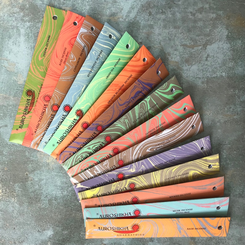 May include: A fan of colorful incense sticks in various scents, including cedar, rose, cinnamon, and jasmine. Each package features a unique marbled design and the brand name "Auroshikha". The incense sticks are arranged on a textured gray surface.