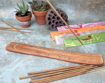 May include: A wooden incense burner with a crescent moon and stars design. The burner is brown with gold accents. There are several incense sticks on the burner and a bundle of incense sticks on the table. The incense sticks are brown and have a light brown scent.