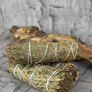 Könnte beinhalten: Zwei Bündel getrockneter grüner Kräuter, fest mit weißem Faden umwickelt. Die Bündel sind übereinander gestapelt, mit einem Holzstück im Hintergrund. Die Kräuter haben ein natürliches, erdiges Aussehen.