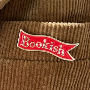 May include: A red enamel pin shaped like a waving banner with the word Bookish written in silver lettering. The pin is attached to a brown corduroy fabric surface with visible vertical ridges and a pocket seam detail.