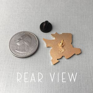 May include: Gold-colored enamel pin featuring a cartoon-style character with a large head and a small body. The pin has a black rubber clutch back and the text "AJB" is visible on the back.