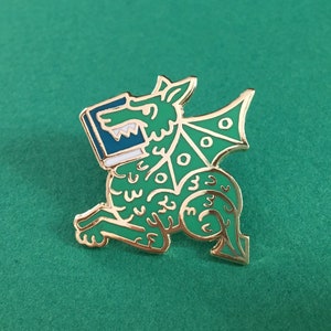 May include: A green and gold enamel pin of a dragon holding a book in its mouth. The dragon has a white and blue book in its mouth and a gold outline.