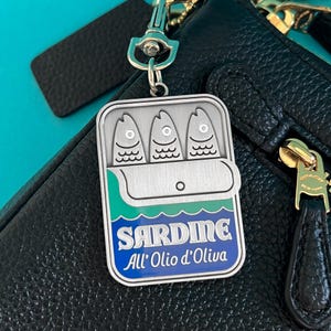 May include: Silver-tone sardine can keychain with three fish inside. The can has a blue and green label with the word "Sardine" and the text "All' Olio d'Oliva". The keychain is attached to a black leather bag.