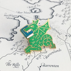 May include: A gold-colored enamel pin featuring a green dragon holding a blue book. The dragon has a white outline and gold accents. The pin is on a white background with a map.