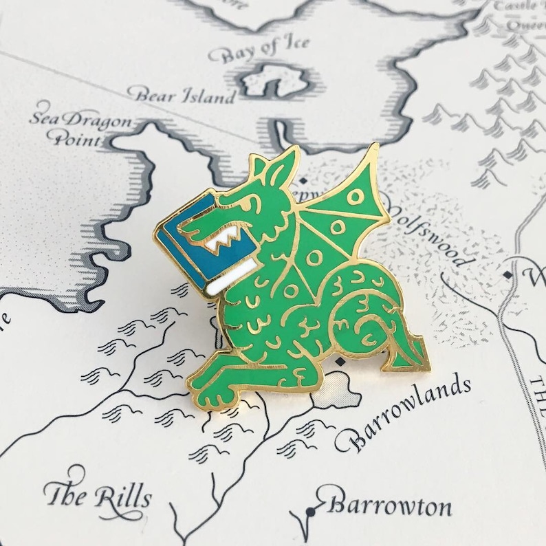 May include: A gold-colored enamel pin featuring a green dragon holding a blue book. The dragon has a white outline and gold accents. The pin is on a white background with a map.