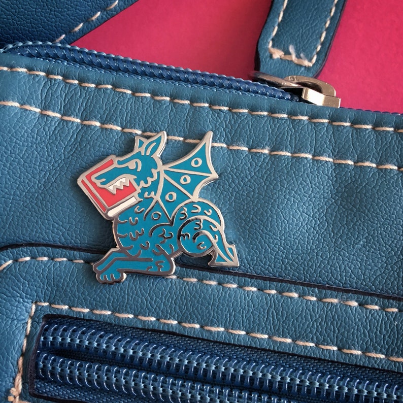 May include: A blue enamel pin of a dragon holding a red book. The dragon has a silver outline and is set against a blue leather background.