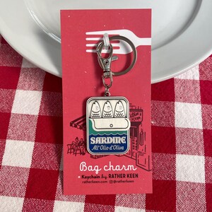 May include: A silver-colored sardine bag charm keychain with a lobster clasp. The charm features a sardine can design with the text "Sardine All'Olio d'Oliva" and the words "Bag charm" below. The background is red.